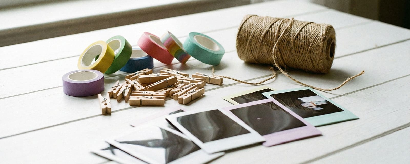 A creative workspace table with rolls of colorful washi tape, a pile of mini wooden pegs, a spool of twine, and a few instant photos scattered on a white surface.