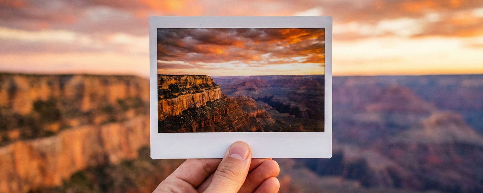 A hand holding a wide-format instant photo up against a real blurred background of a stunning sunset over a canyon